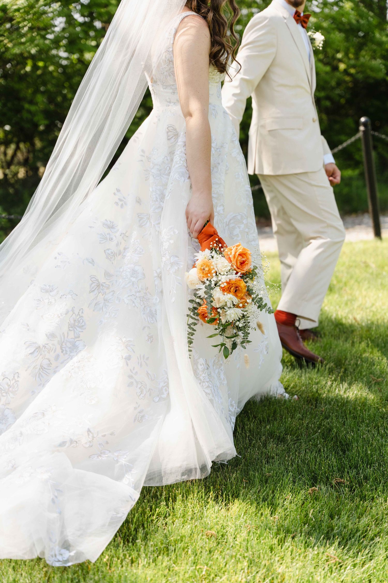 Bride in floral gown with veil holds orange bouquet walking beside groom in tan suit on green lawn.