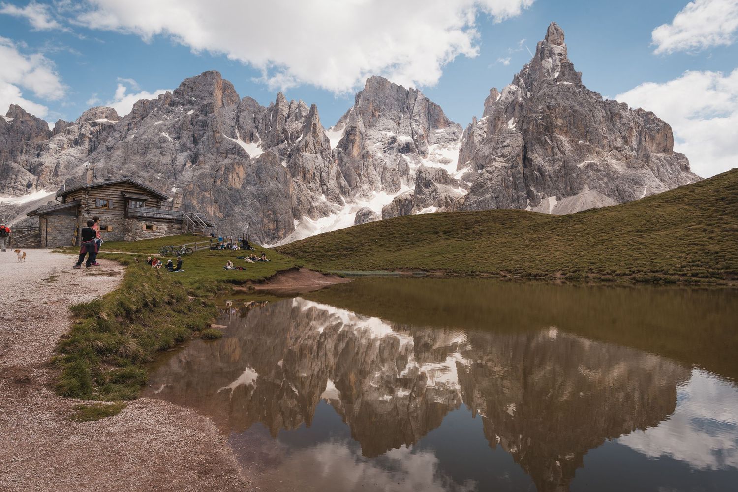 Baita di montagna e il profilo maestoso delle Pale di San Martino che si specchiano sulle acque limpide di un laghetto alpino sotto un cielo
