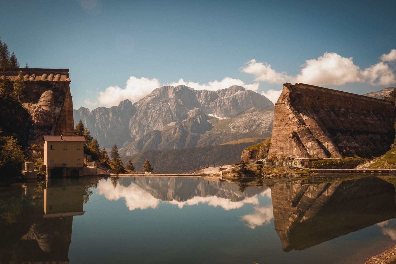 Vista panoramica dei resti della diga del Gleno che si specchiano nelle acque calme del lago, con le vette alpine e un cielo azzurro