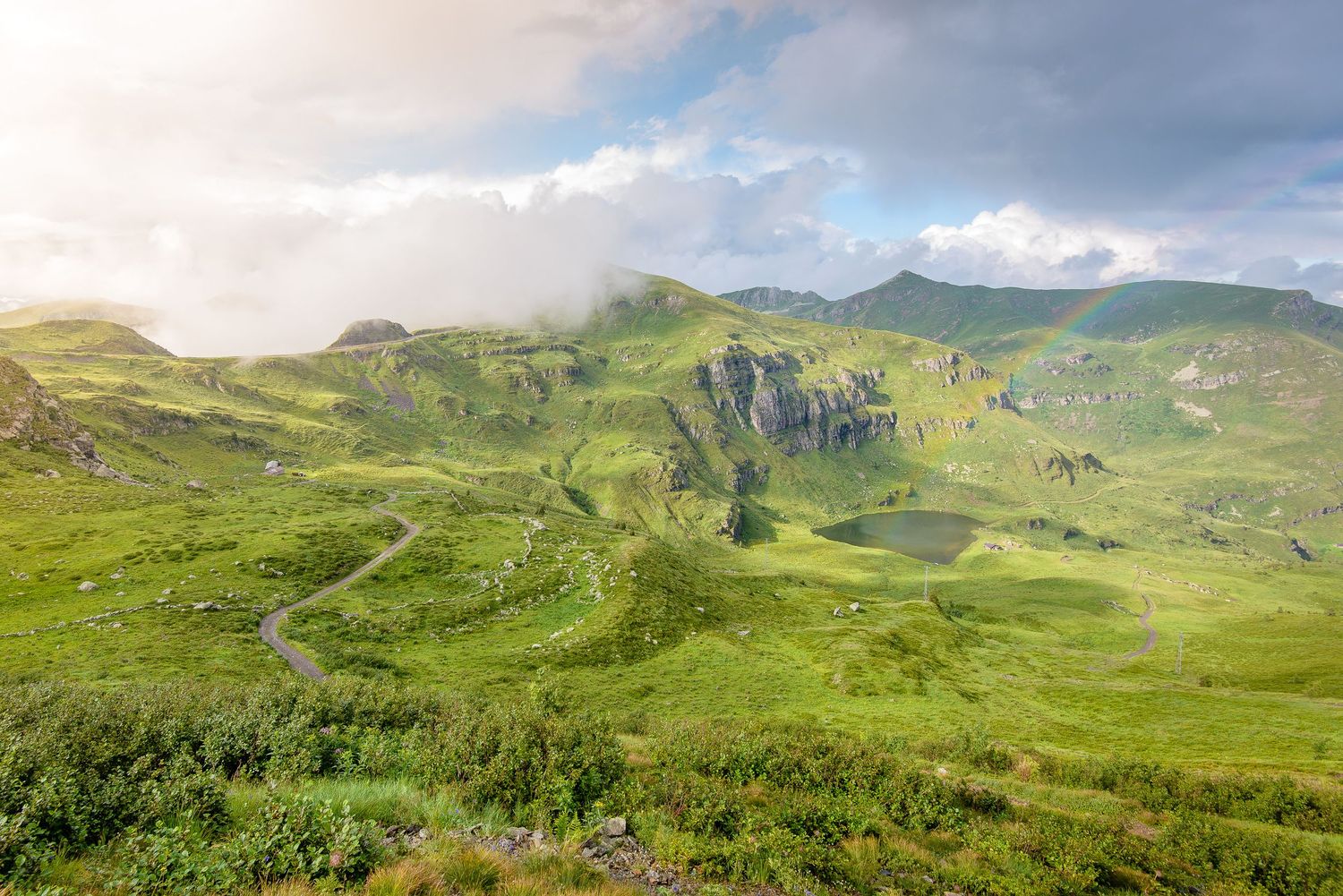 Vista dall'alto di un piccolo lago alpino incastonato tra verdi pascoli di montagna, con un accenno di arcobaleno e un sentiero che serpeggi