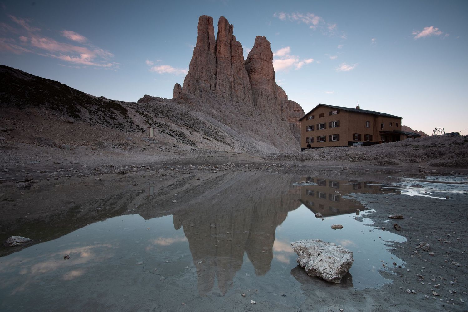 Le imponenti Torri del Vajolet e il Rifugio Re Alberto I che si specchiano in una pozza d'acqua sulla conca rocciosa di giorno.