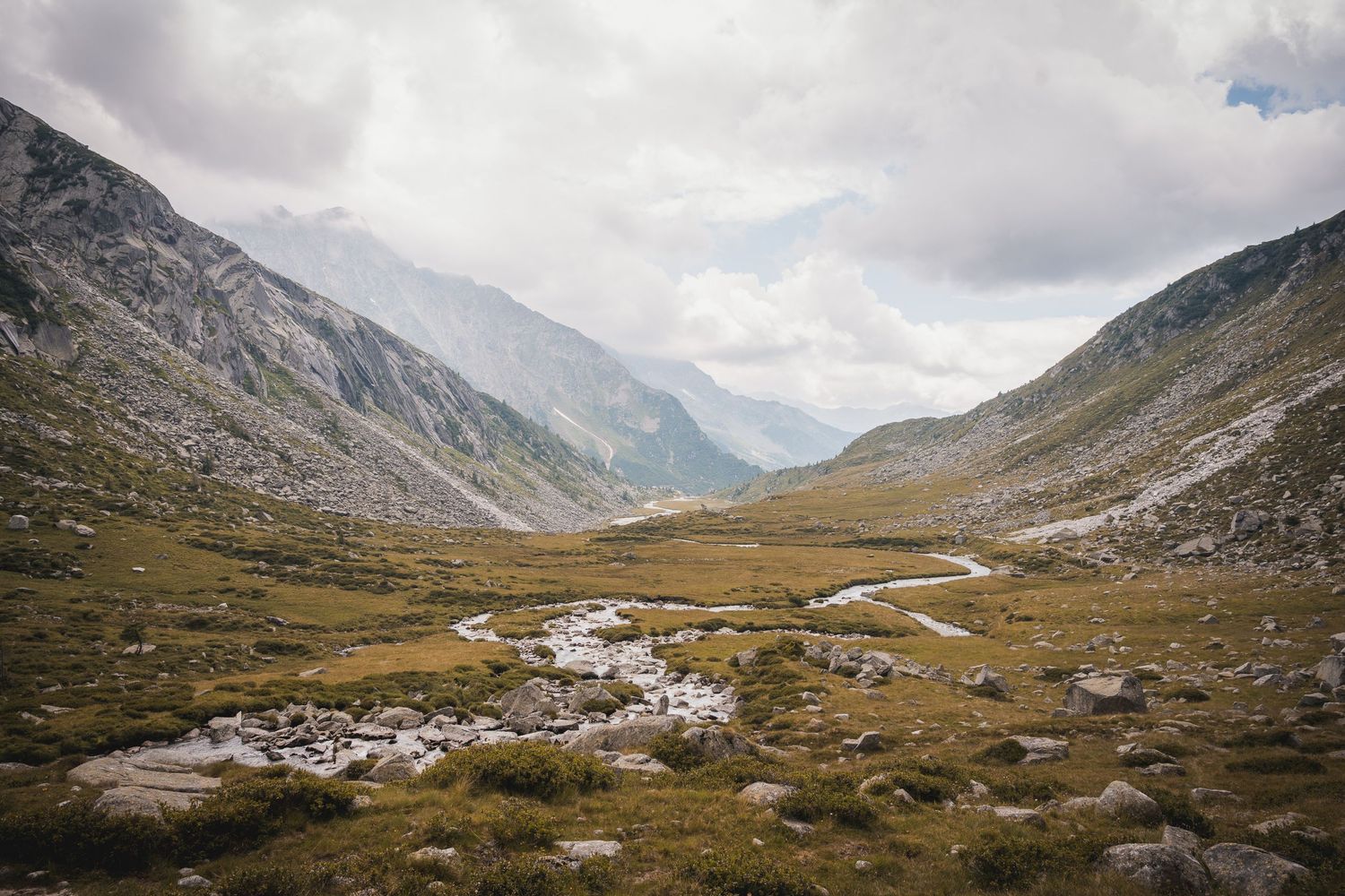 Ampia vista panoramica della piana della Val Adamé con un torrente che forma meandri tra i prati dorati, racchiusa da imponenti versanti