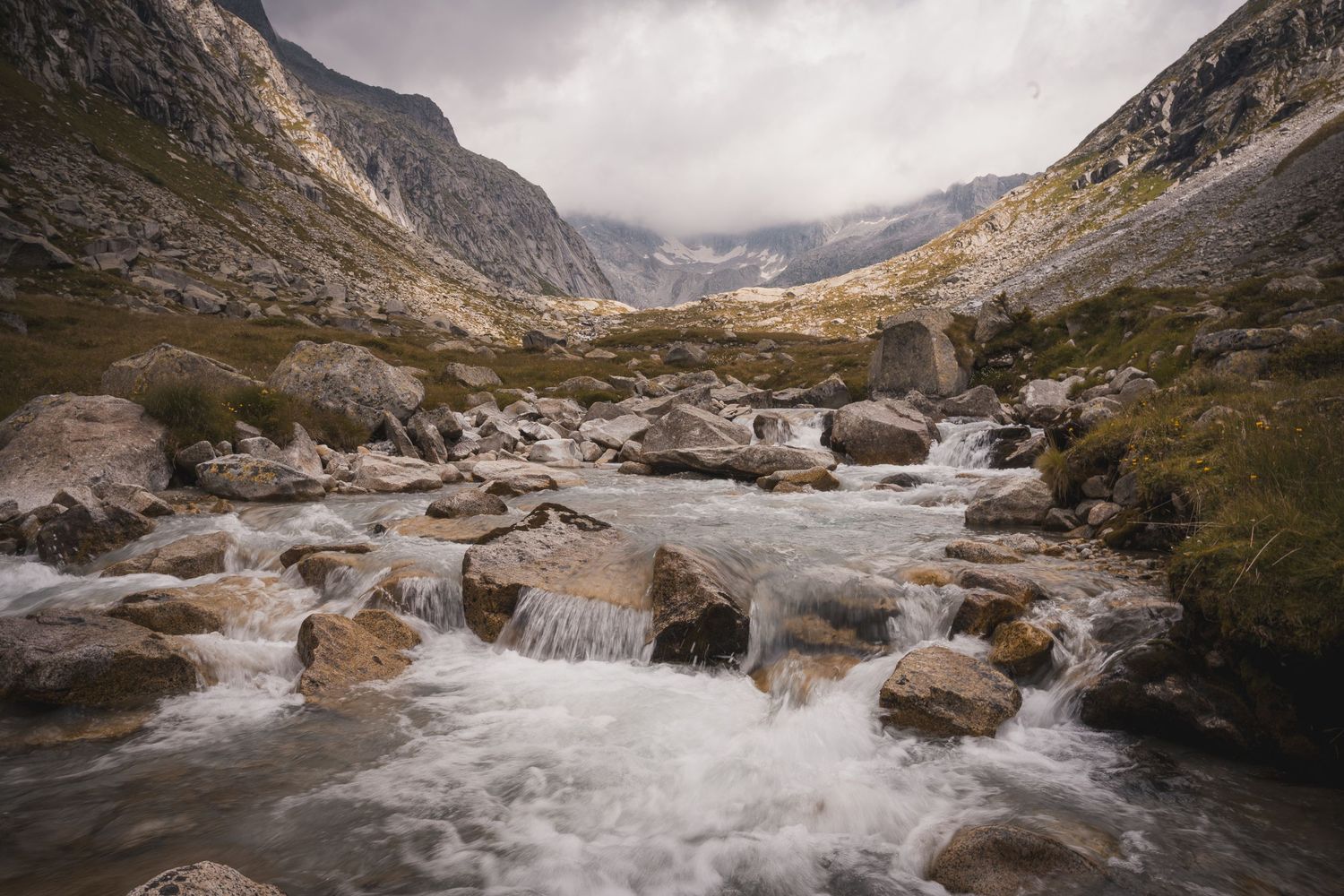 Un torrente montano impetuoso scorre tra rocce in primo piano, all'interno di una valle alpina selvaggia dominata da vette rocciose e cielo