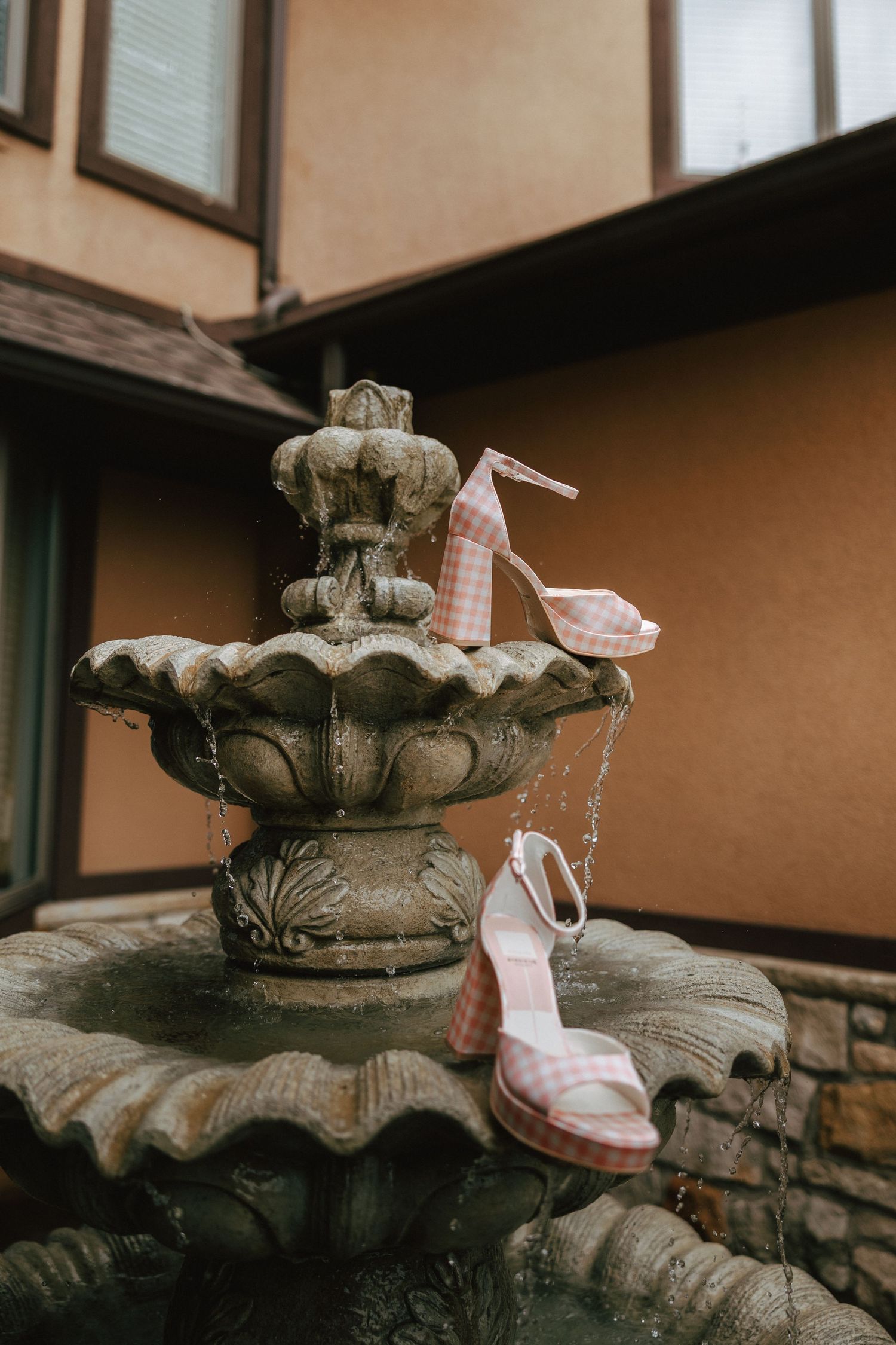Pink gingham heeled sandals displayed on a stone tiered garden fountain outside a building.