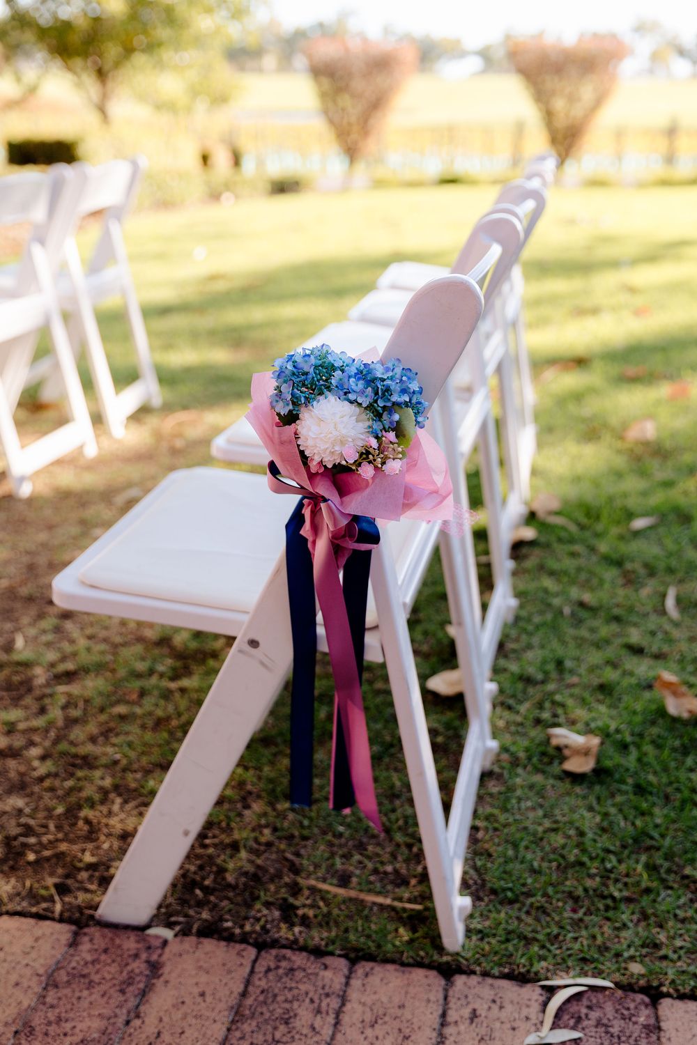 White folding chairs decorated with pink and blue floral bouquets tied with ribbons for an outdoor wedding ceremony.