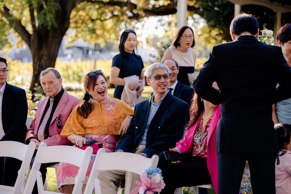 Guests laughing and smiling during an outdoor wedding ceremony seated on white chairs in a sunny garden setting.