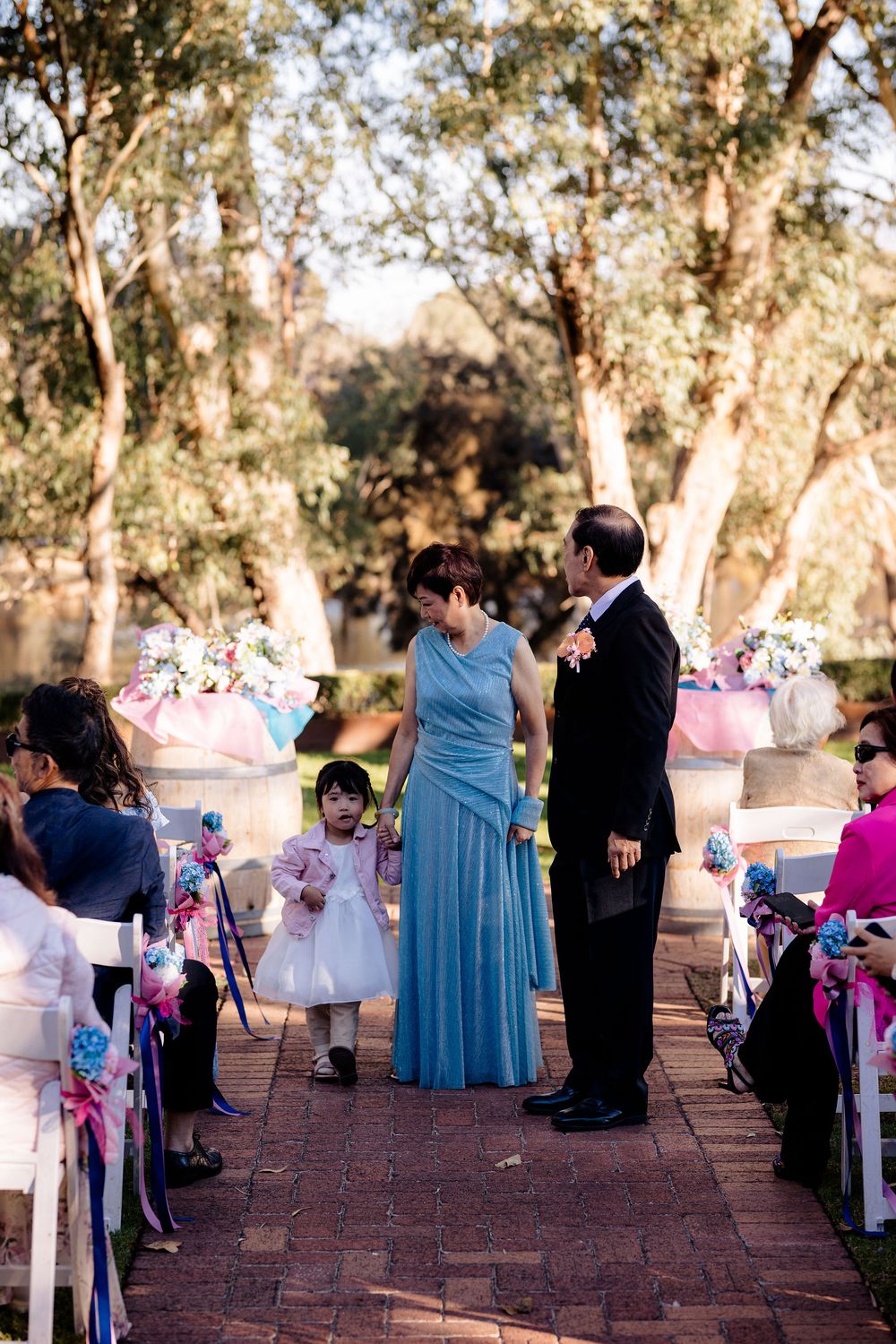 A woman in a blue gown and man in a tuxedo stand with a young girl in a white dress during an outdoor wedding ceremony.