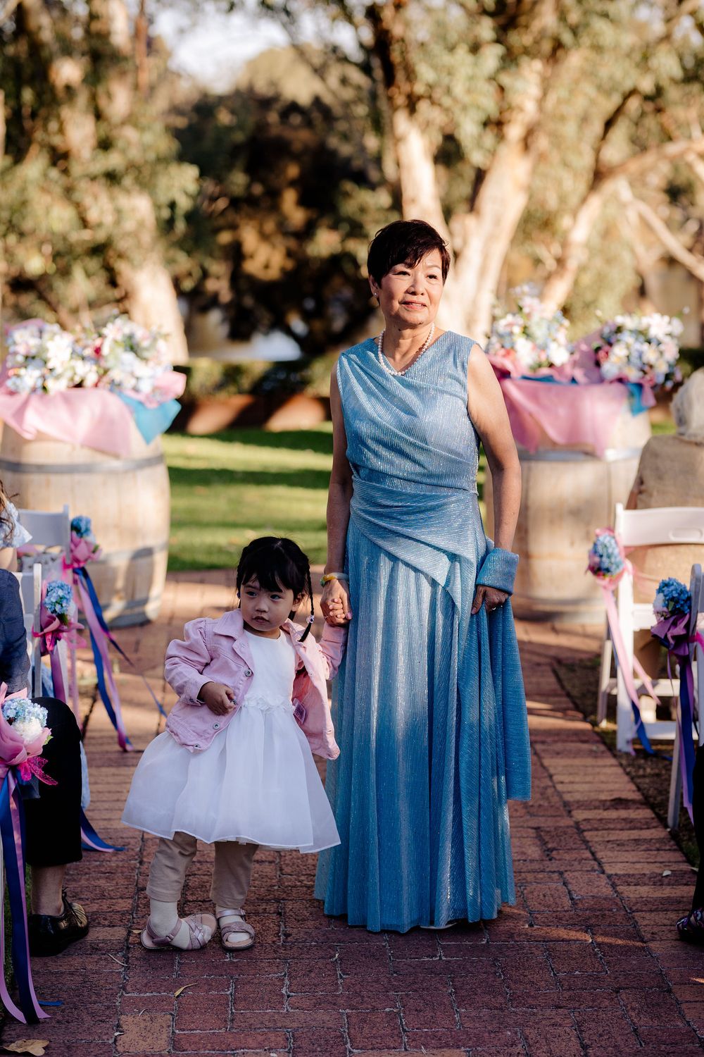 Woman in blue gown and young girl in white dress walk down outdoor wedding aisle with floral decorations.
