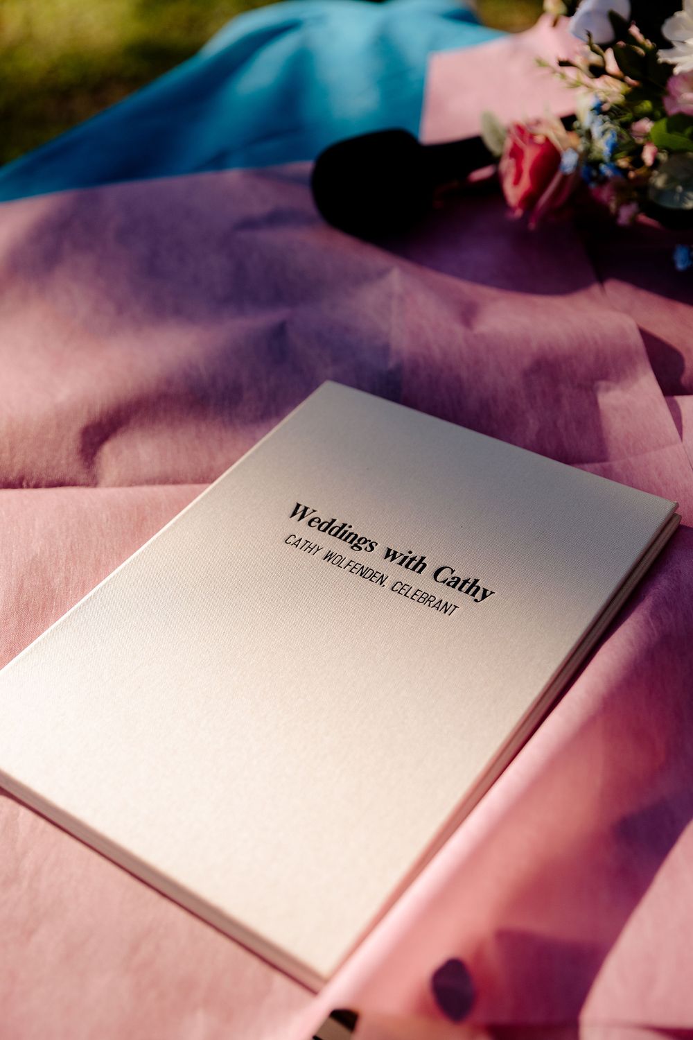 Wedding ceremony booklet titled Weddings with Cathy, celebrant, resting on pink fabric with flowers nearby.
