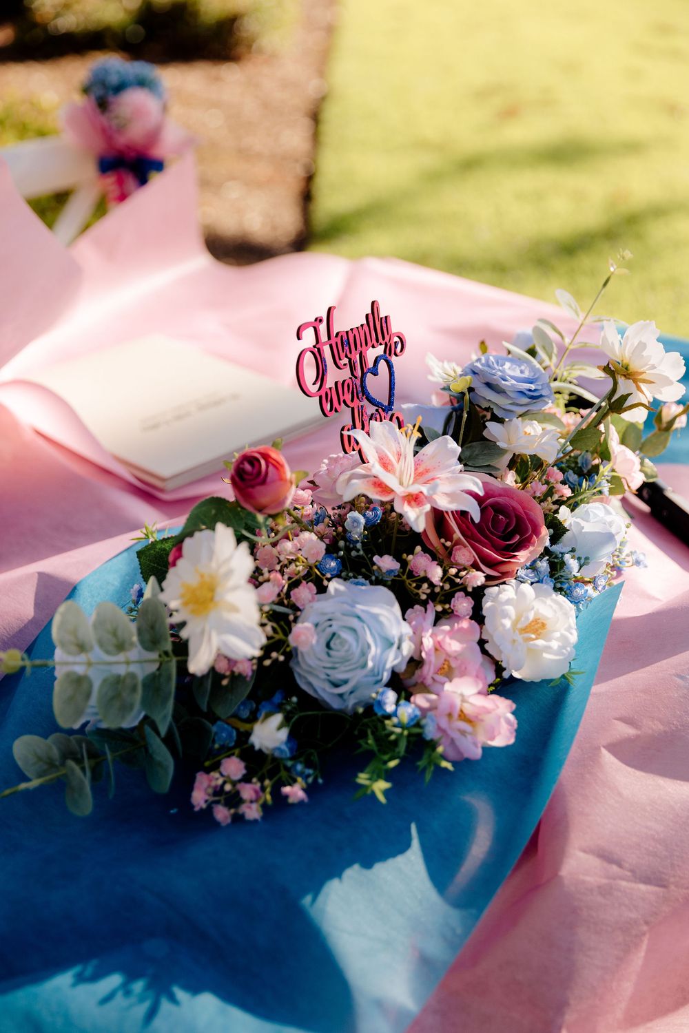 Elegant wedding floral centerpiece with pink and blue roses, daisies, and a Happily Ever After sign on a decorated table.