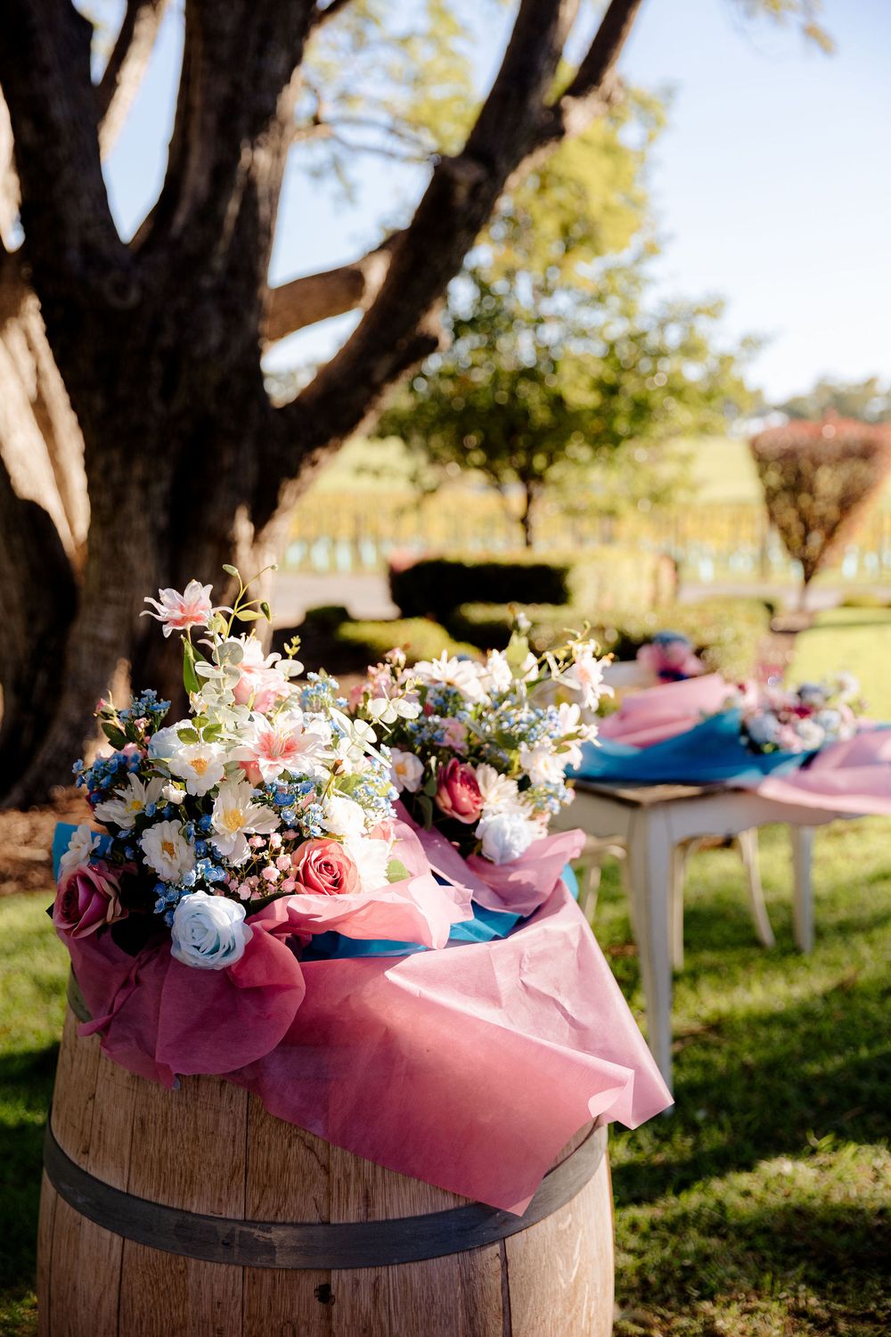 Rustic wooden barrels decorated with pink and blue floral arrangements for an outdoor garden wedding ceremony.