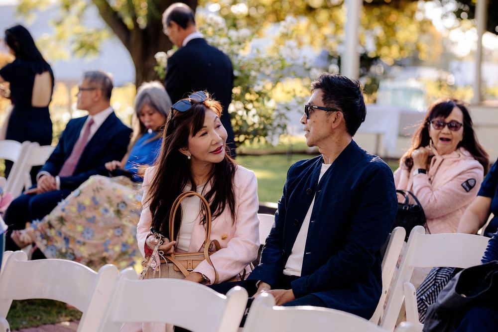 Guests seated in white chairs at an outdoor wedding ceremony, smiling and conversing in elegant attire.