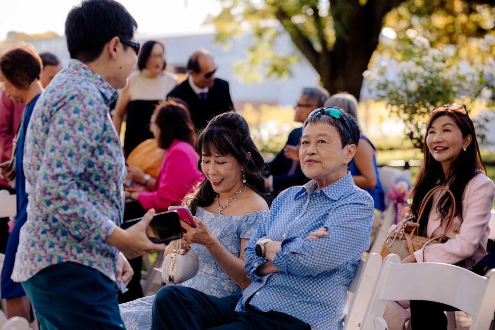 Guests seated outdoors at a wedding ceremony, smiling and conversing in a sunlit garden setting.
