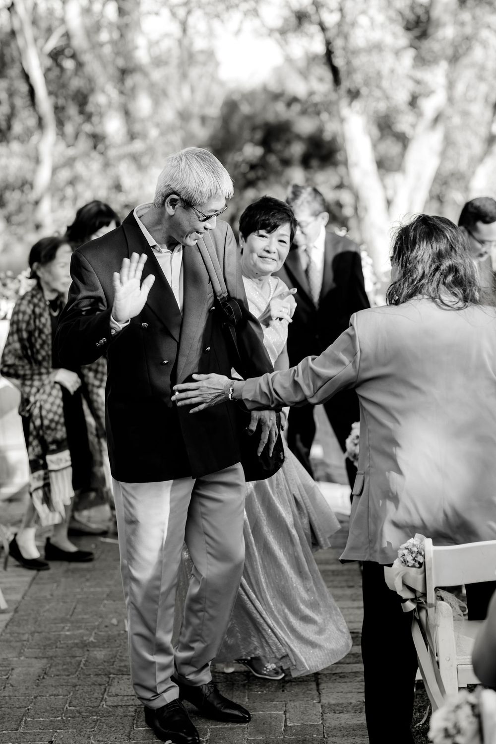 Black and white photo of elderly couple greeting guests at an outdoor wedding ceremony on a brick pathway.