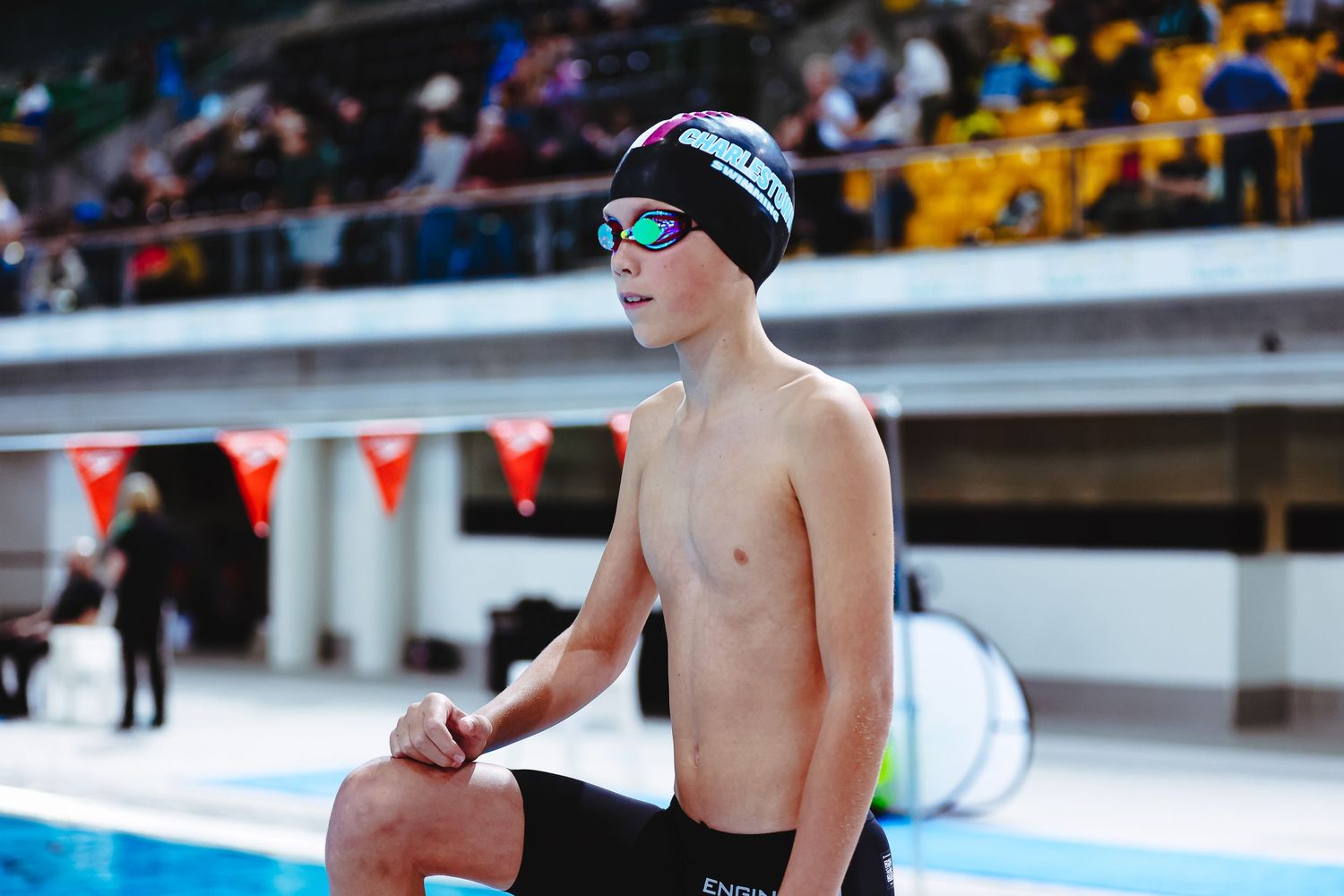 Young male swimmer in orange cap and goggles stands poolside in red swimwear at an indoor aquatics competition.