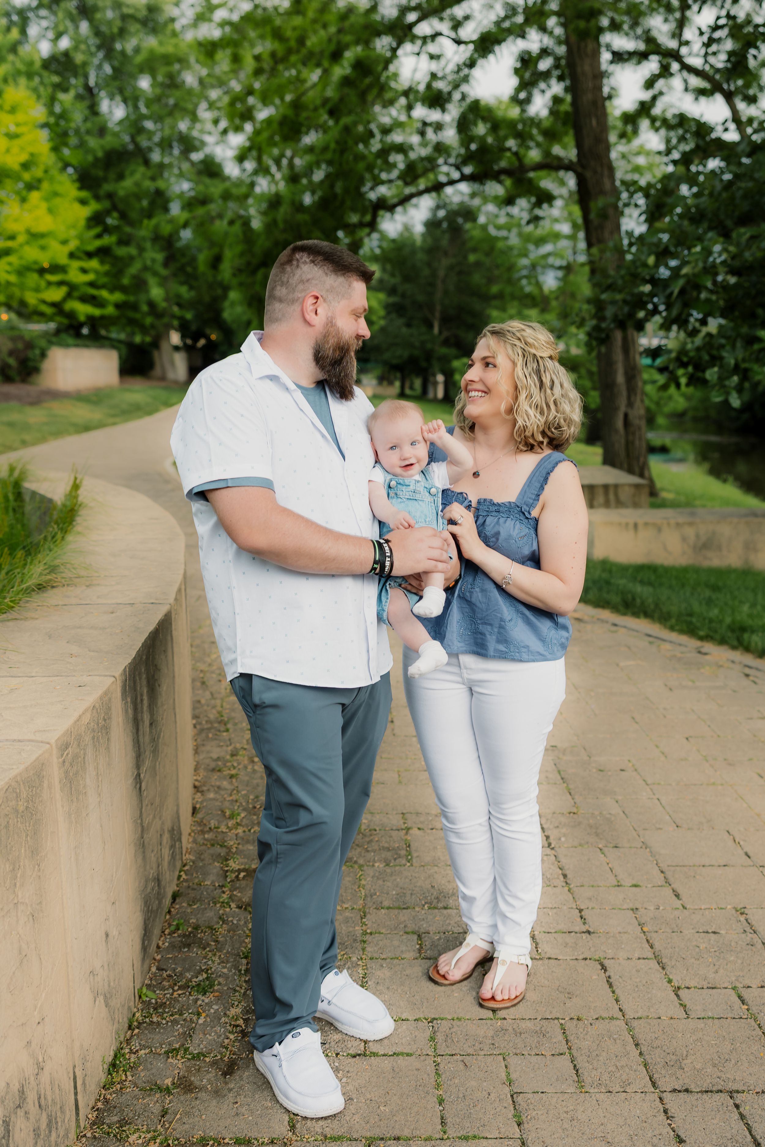 A happy couple smiles at each other while holding their baby outdoors in a lush green park setting.