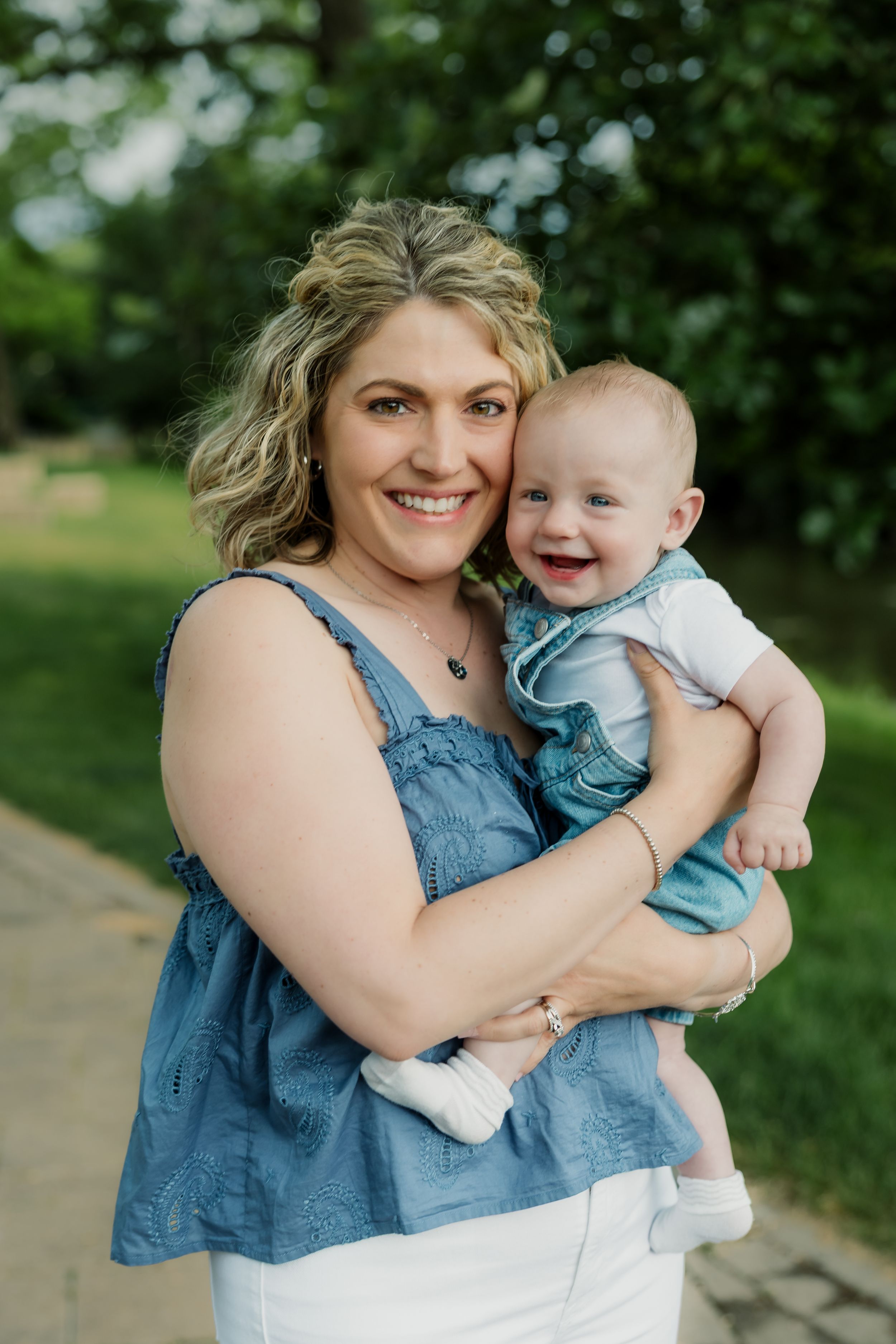 Smiling woman in blue top holding a happy baby in denim outfit outdoors in a green park setting.
