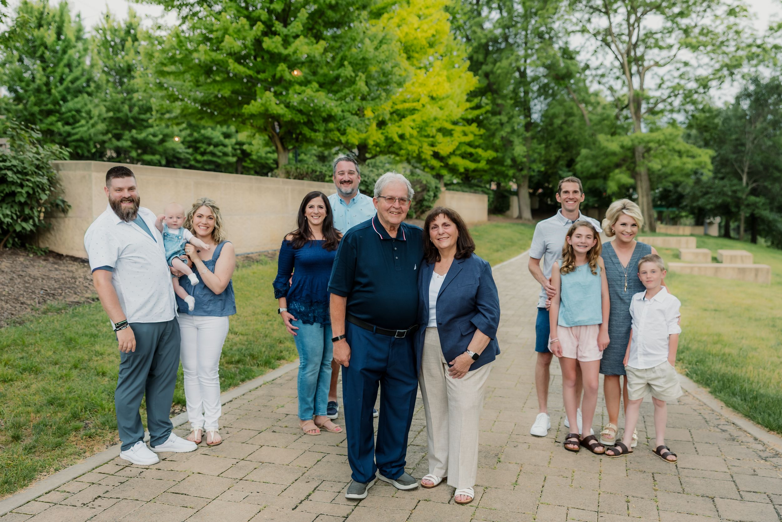 Multi-generational family portrait outdoors on a paved path surrounded by lush green trees and garden landscaping.