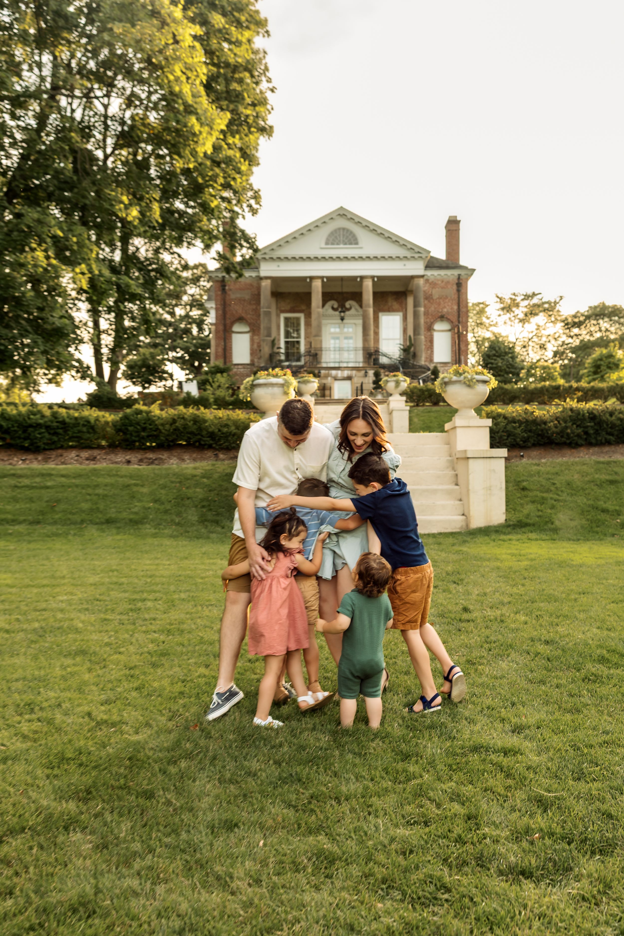 Family of six sharing a warm group hug on green lawn before a classic brick colonial mansion at golden hour.
