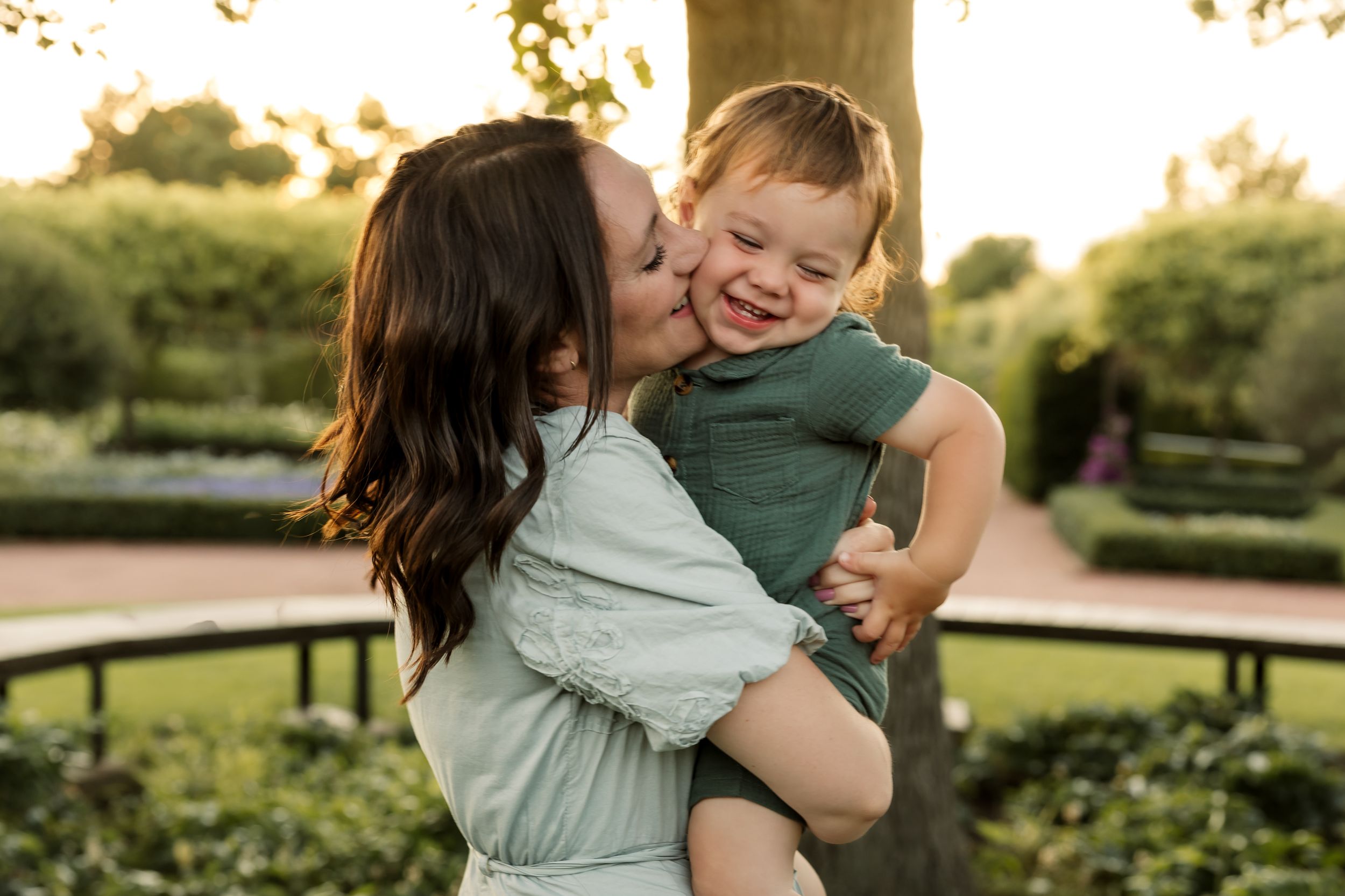 A mother lovingly kisses her laughing toddler son outdoors in a lush garden during golden hour.