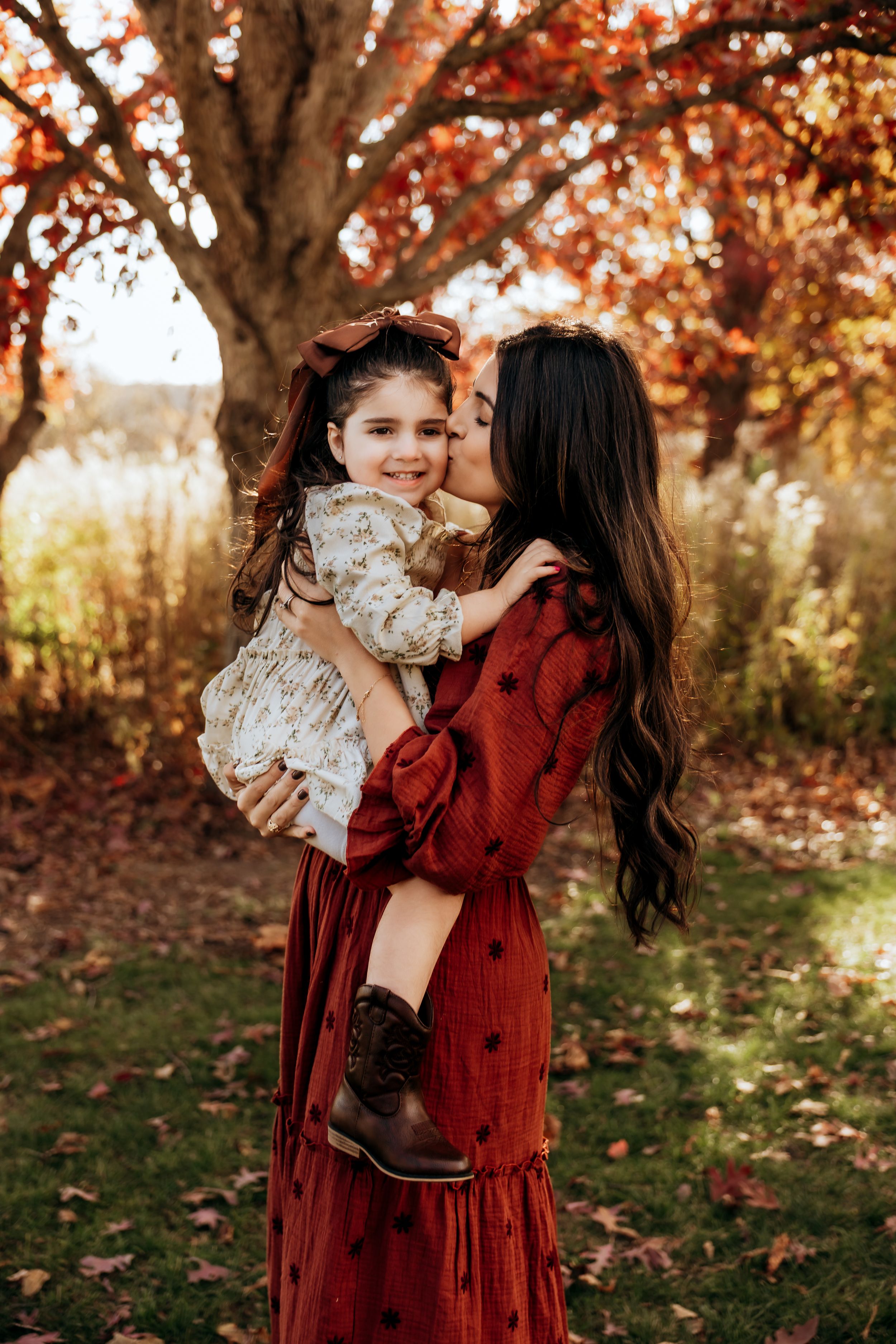 A mother in a red dress kisses her smiling daughter outdoors among vibrant autumn foliage.