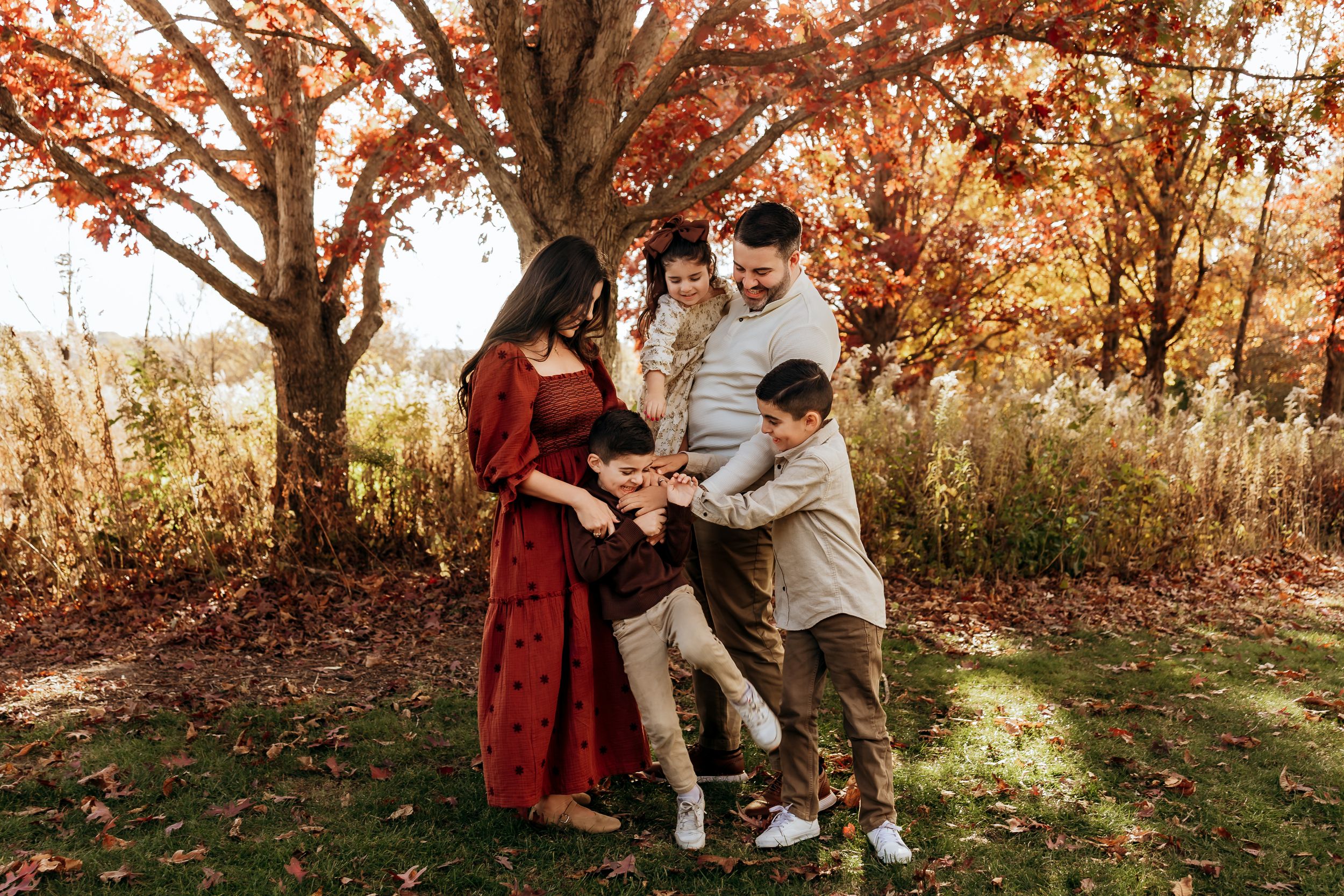 Family of four enjoying autumn outdoors under vibrant red foliage, parents and two children laughing together on green grass.