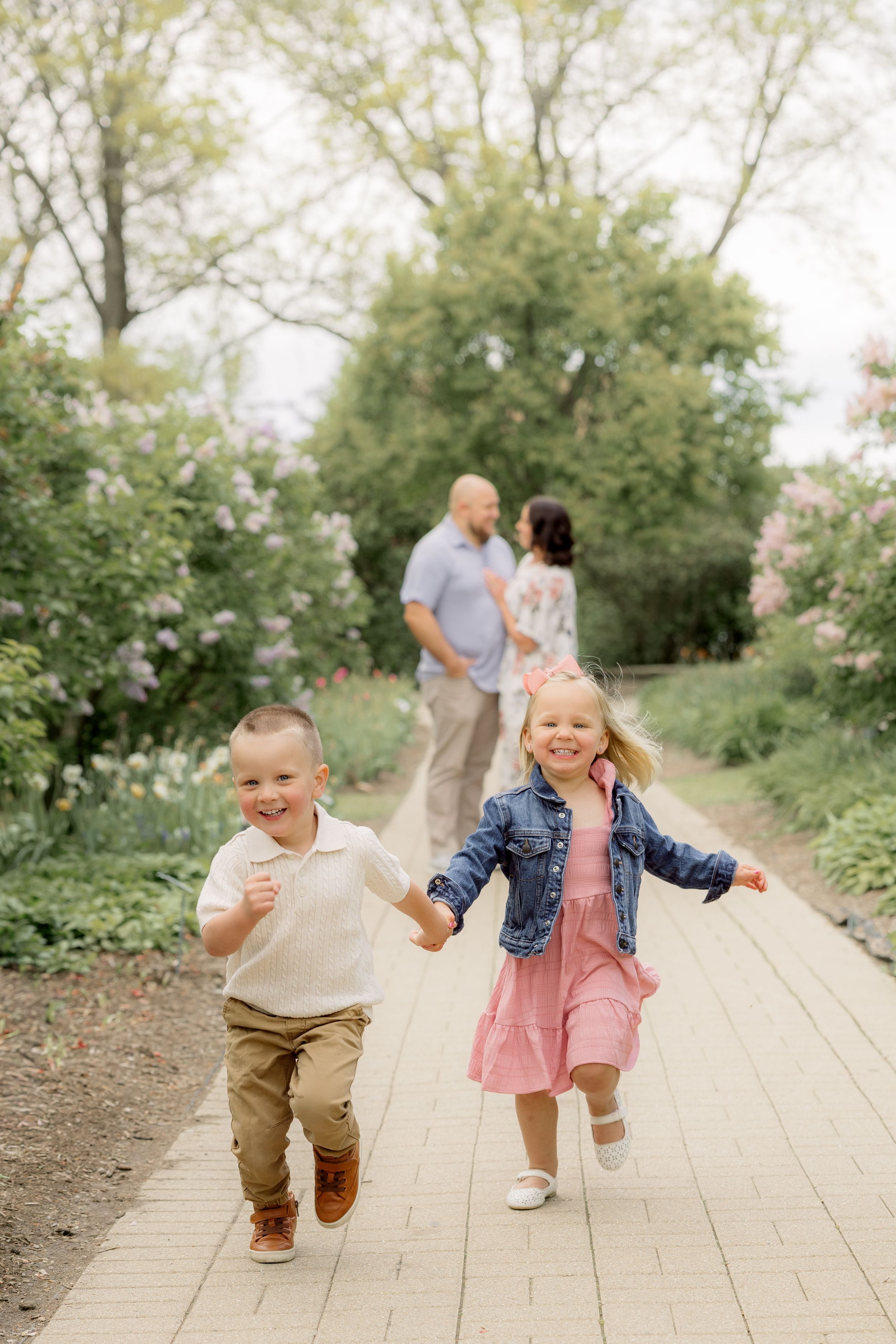 Two young children run joyfully down a garden path while their parents share a moment together among blooming trees.