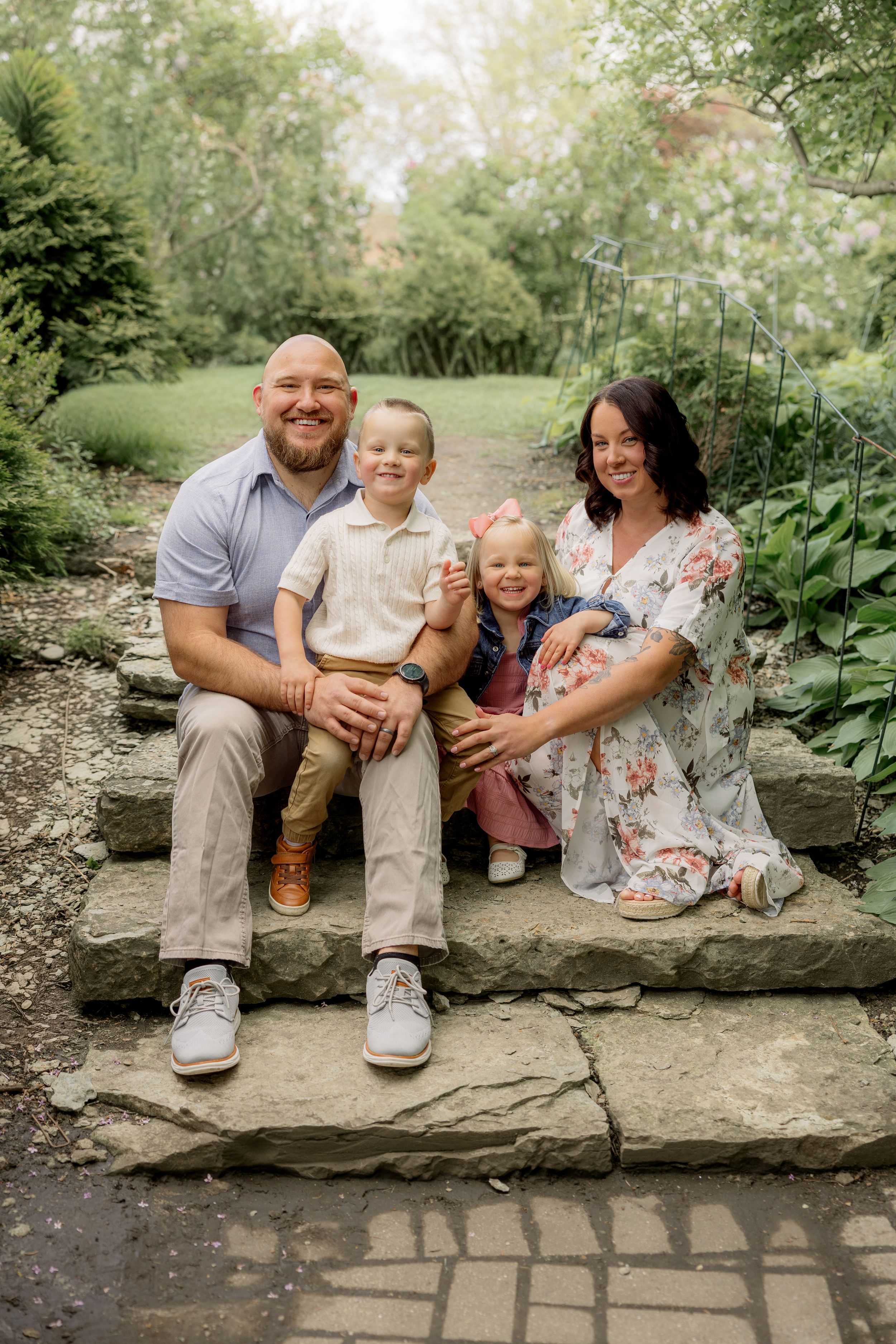 Family of four smiling on stone steps in a lush garden, parents with two young children in coordinated outfits.
