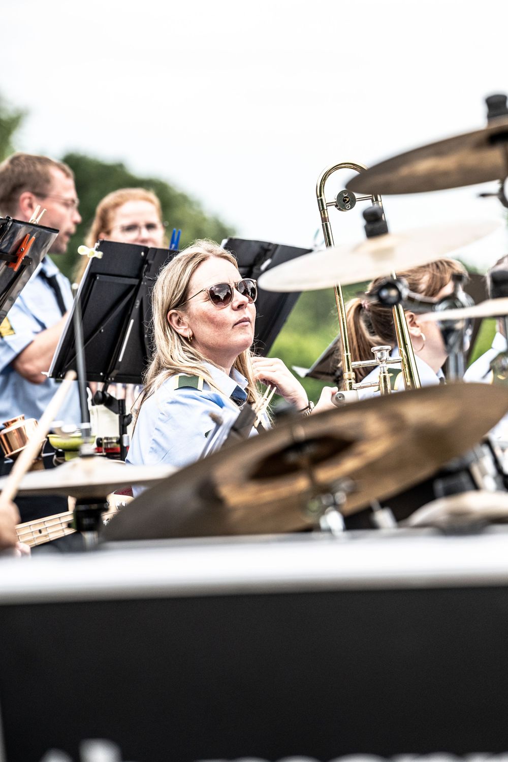 Blonde woman in sunglasses plays drums in an outdoor brass band performance with musicians and music stands visible.