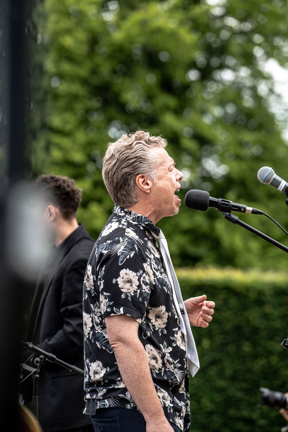 Middle-aged man in floral shirt singing at outdoor microphone stand with green trees in background.