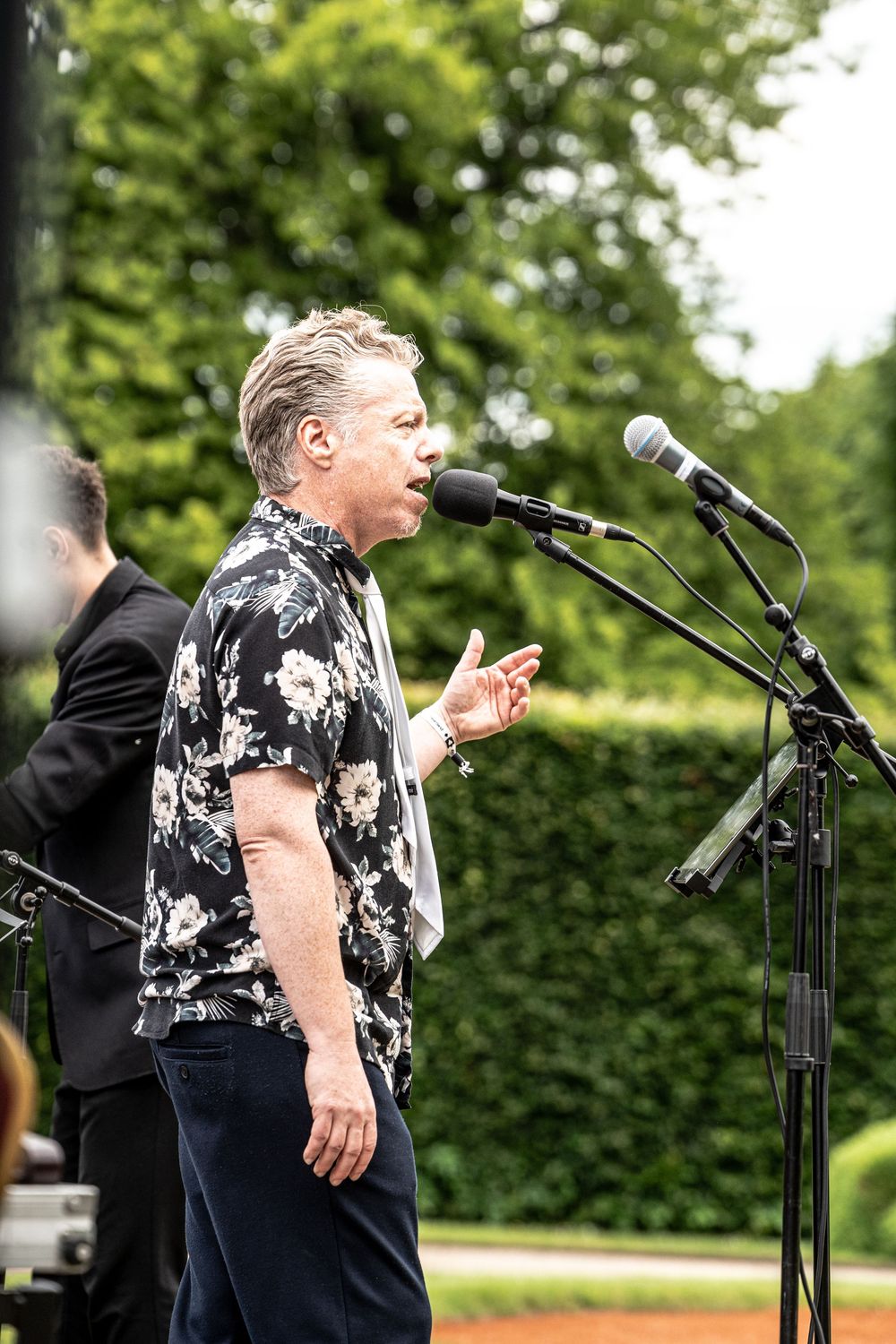 Man in floral shirt singing at outdoor microphone stand with green trees in background.