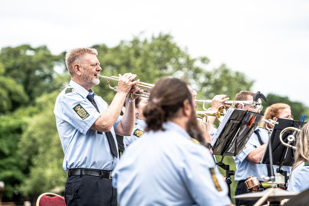 Police band musicians in uniform playing trumpets outdoors at a summer concert surrounded by lush green trees.