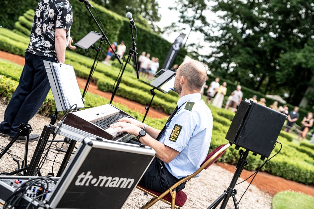 Police officer playing keyboard on a Thomann stand at an outdoor music event in a sunny park.