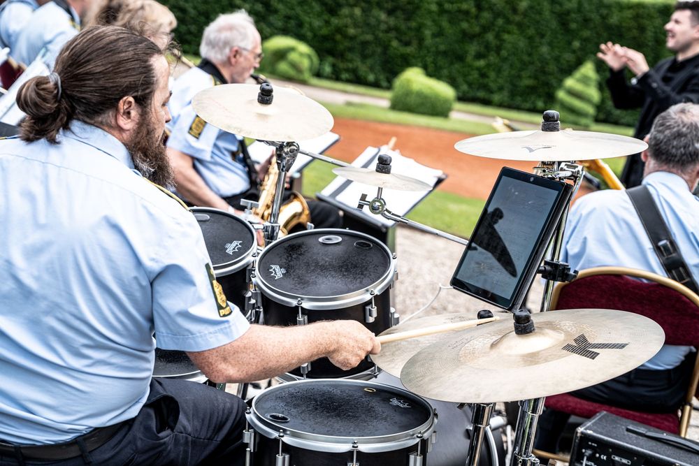 A drummer in a blue uniform plays drums outdoors at a formal band performance, with cymbals and a tablet visible.