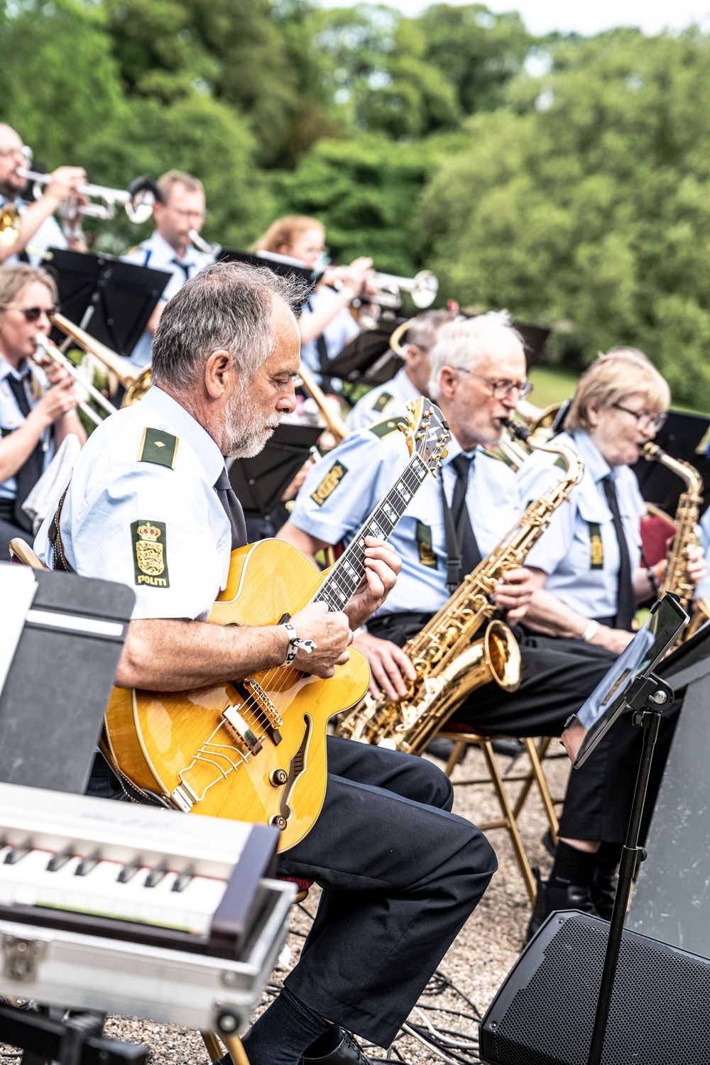 Uniformed police orchestra members perform guitar and saxophone outdoors.