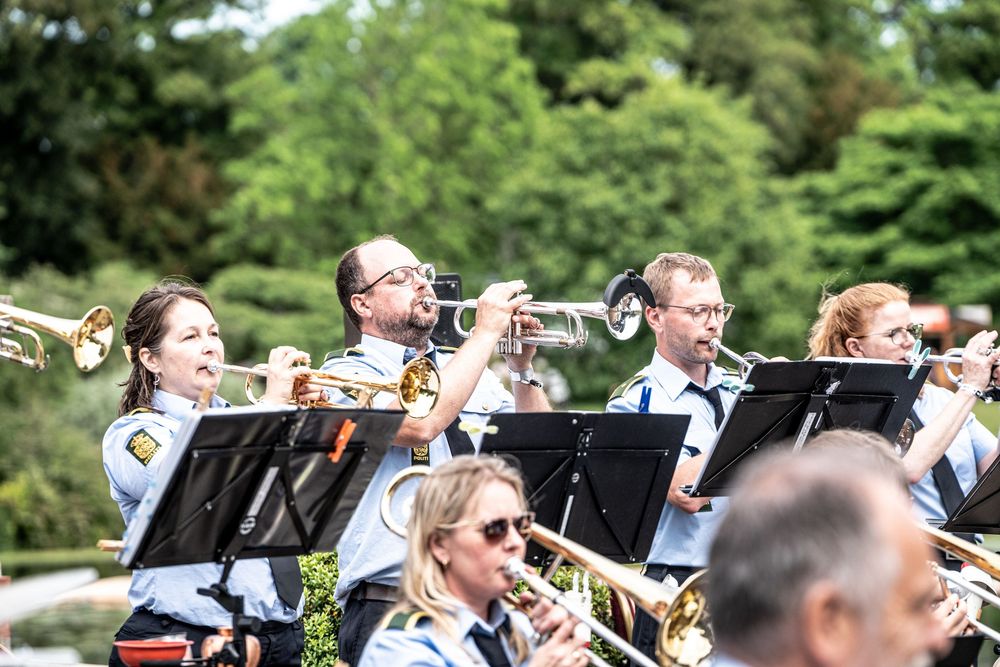 Brass band musicians in uniforms perform outdoors at a summer concert surrounded by lush green trees.