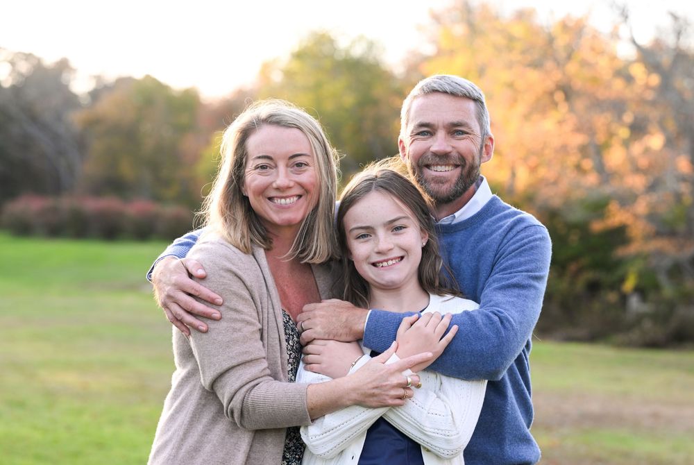 Happy family of four posing outdoors in autumn park, parents smiling with young daughter holding newborn baby.