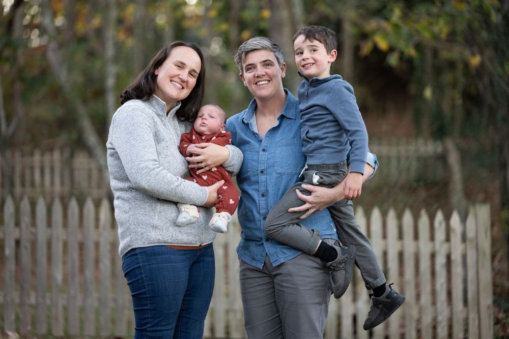 Happy family of four posing outdoors near a white picket fence, parents holding two young children.