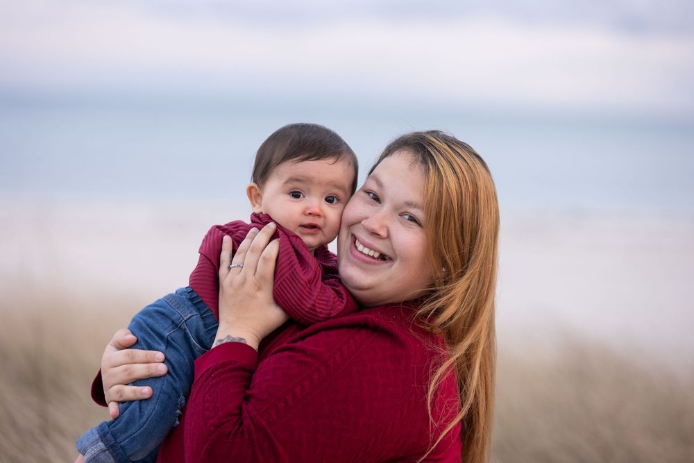 A smiling woman with red hair holds a baby on a sandy beach, both wearing red outfits on a cloudy day.