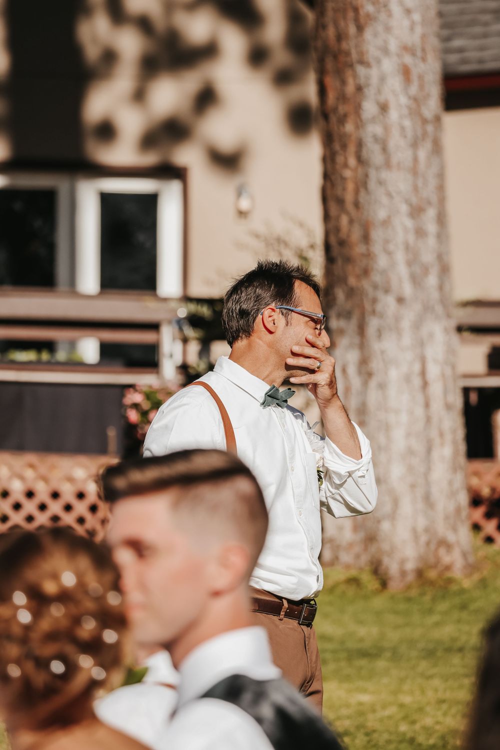 Emotional wedding guest wiping tears at outdoor ceremony, wearing white shirt with suspenders and glasses.