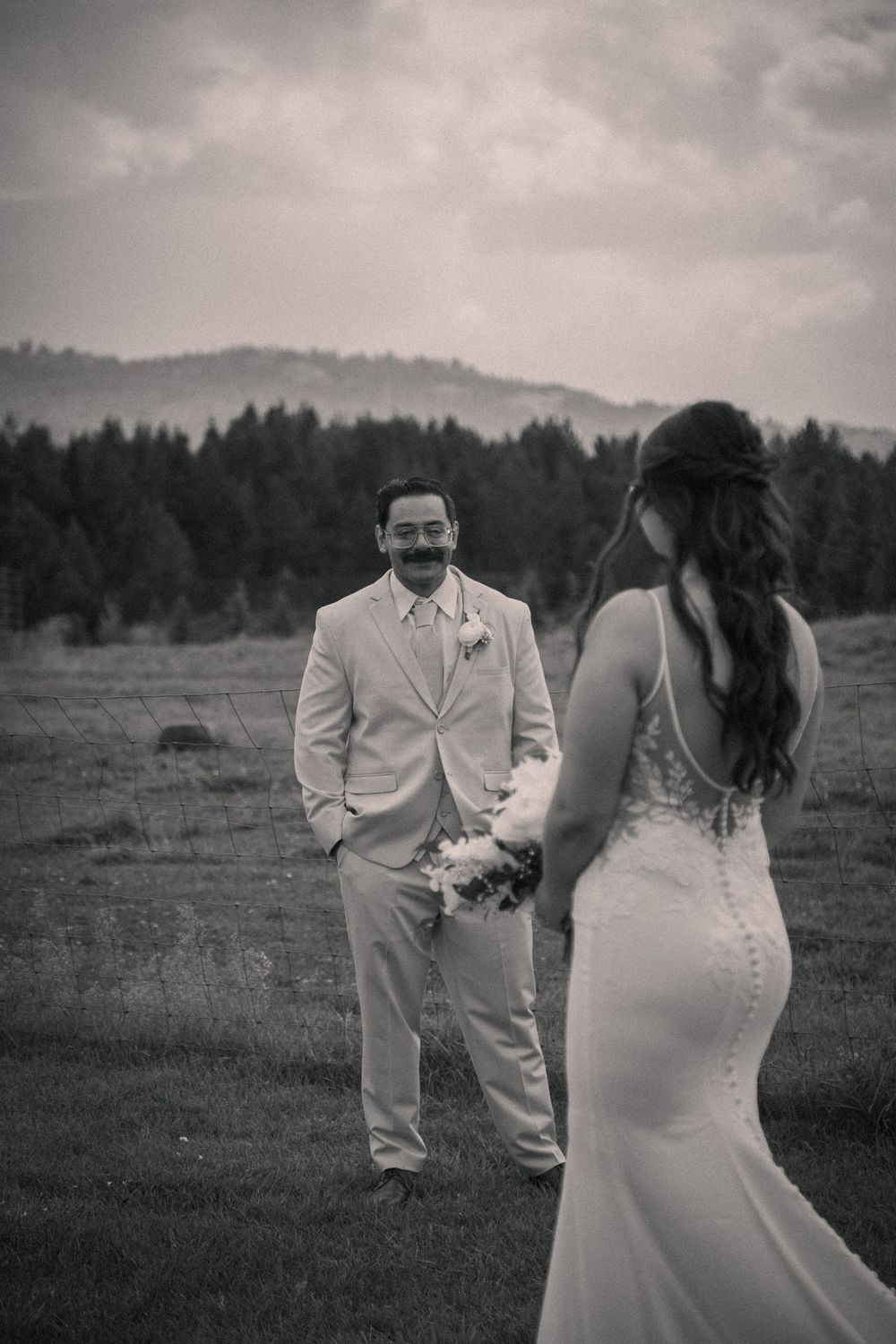Black and white photo of a groom smiling at his bride in a field with pine trees and mountains in the background.