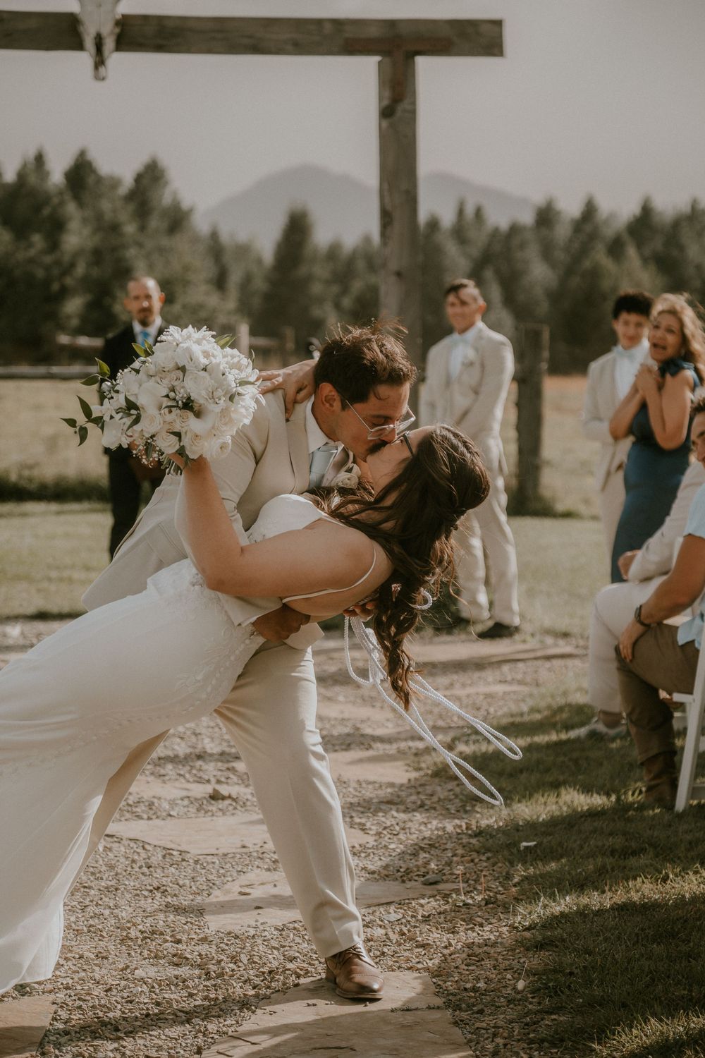 Bride and groom share a romantic dip kiss at outdoor wedding ceremony with mountain backdrop and guests watching.