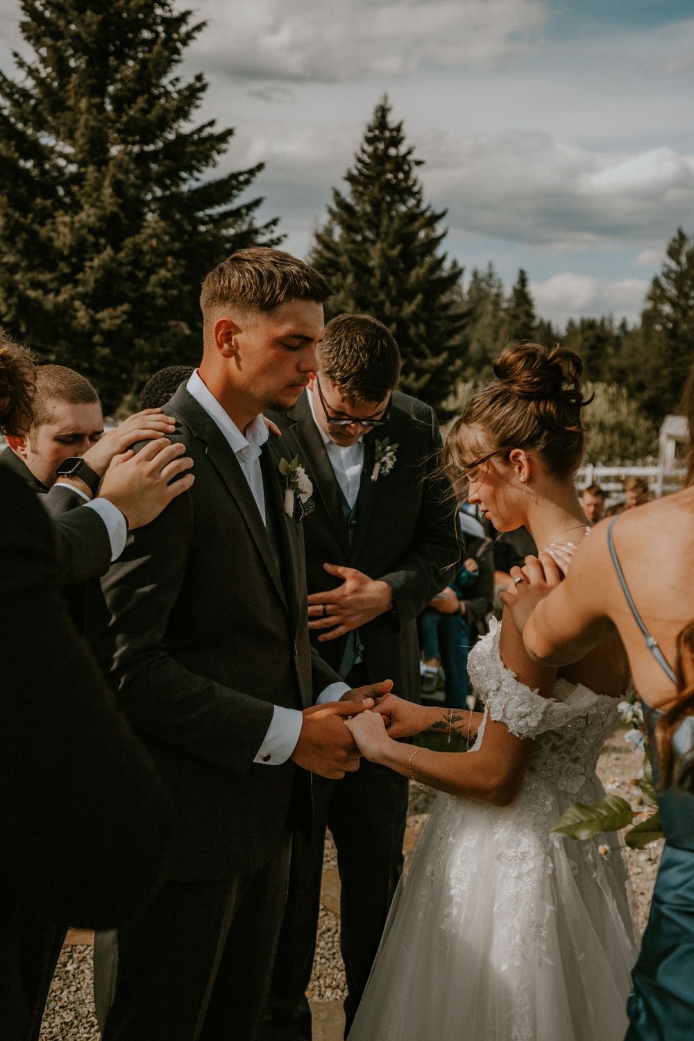 Bride and groom exchange rings outdoors at wedding ceremony surrounded by guests with tall pine trees in background.
