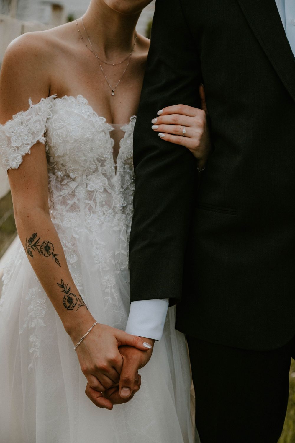 Bride in lace off-shoulder gown holds hands with groom in black suit, showcasing floral arm tattoo and diamond ring.