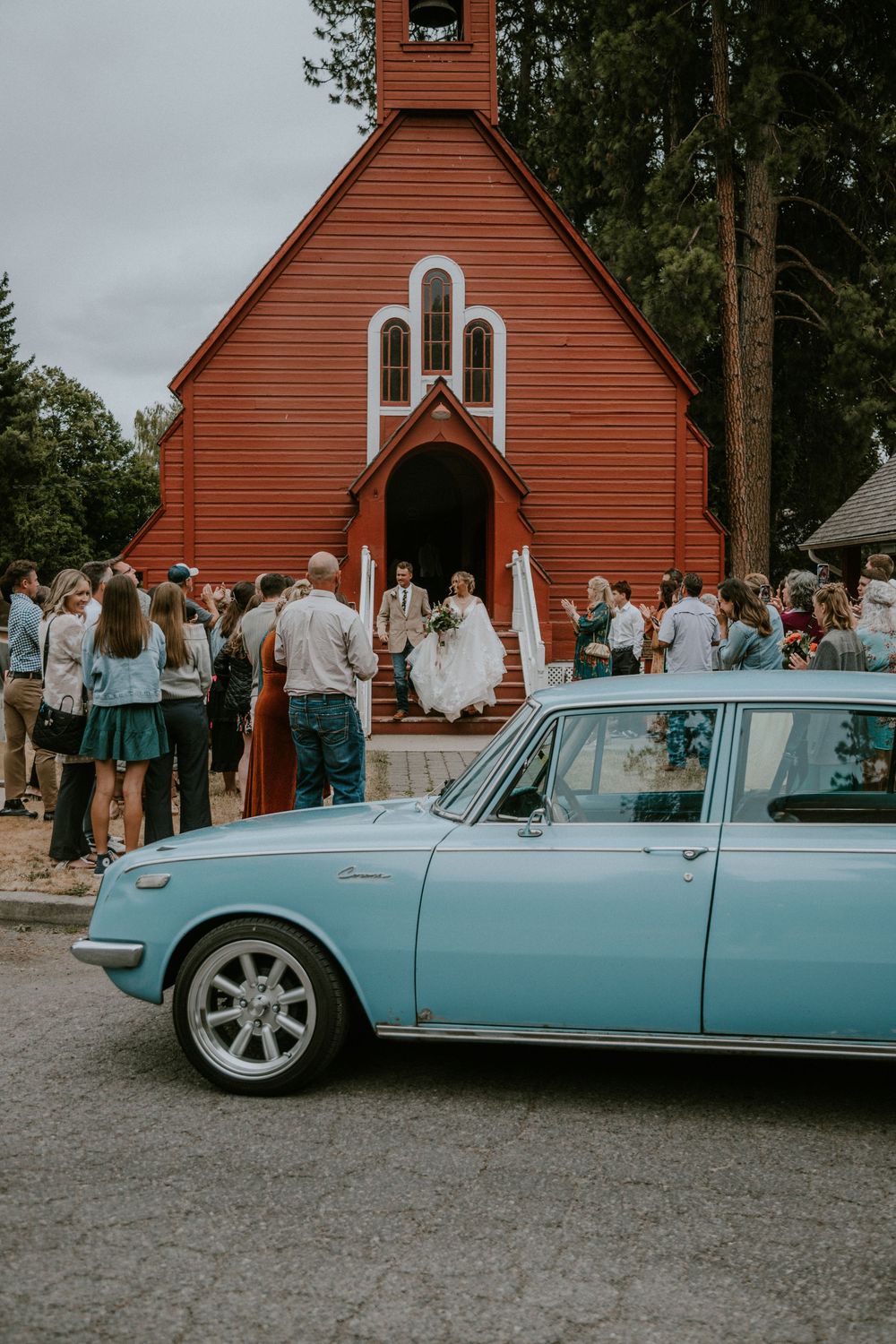 Vintage blue car parked outside a red wooden chapel during a wedding ceremony with guests gathered on the steps.
