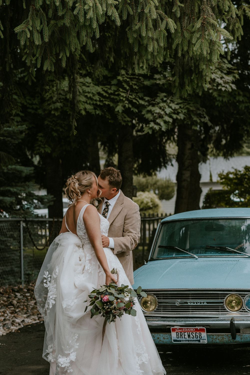 Bride and groom share a romantic moment beside a vintage teal car, bride holding a lush bouquet outdoors.