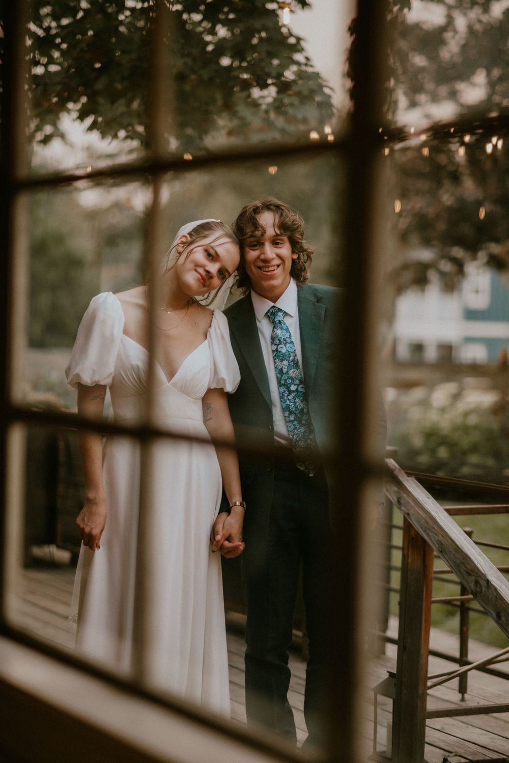 A smiling bride and groom hold hands on a wooden deck, photographed through a window at their outdoor wedding.