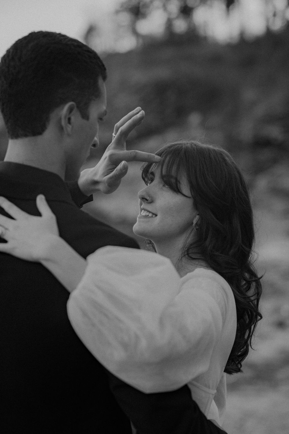 Black and white photo of a smiling bride touching her groom's face while embracing outdoors.
