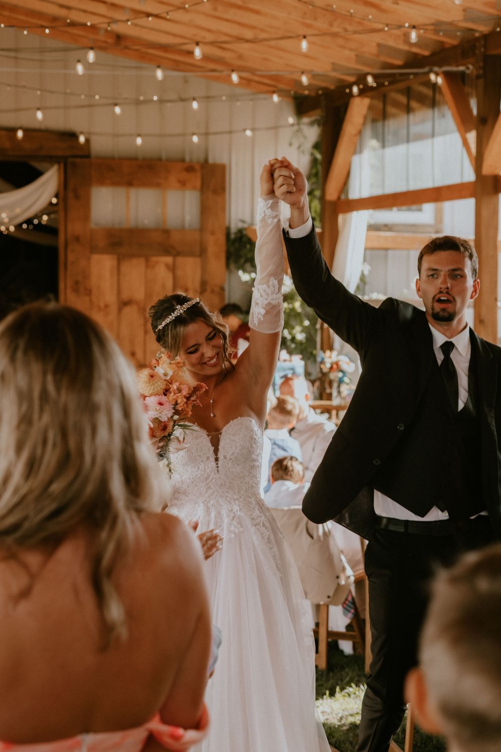 Joyful newlyweds raise hands in celebration at rustic barn wedding ceremony, bride in lace gown holding bouquet.