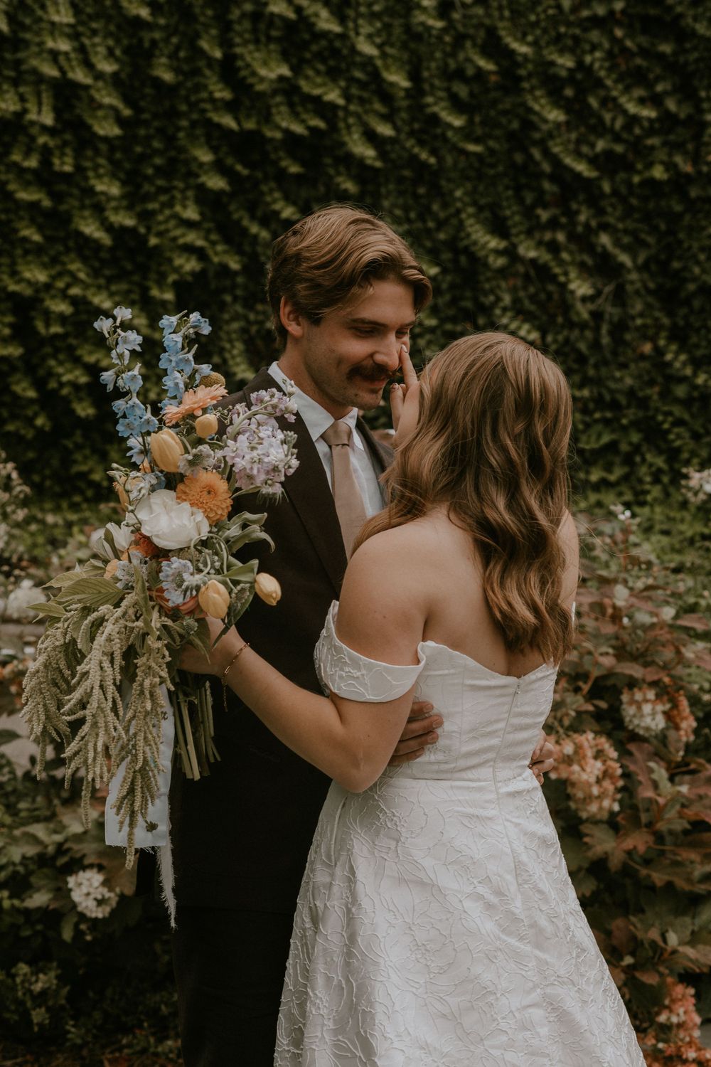 A bride in an off-the-shoulder white gown embraces a groom in a dark suit holding a wildflower bouquet before an ivy wall.
